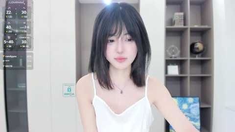 Media: Video of a young Asian woman with straight black hair and fair skin, wearing a white camisole, standing in a modern, minimalist room with a digital display showing weather data.