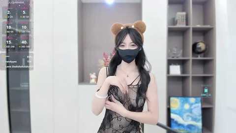 Media: Video of a young Asian woman with long black hair, wearing a black face mask, a brown bear headband, and a sheer black lace dress, standing in a modern, minimalist room with a large digital clock and a bookshelf in the background.