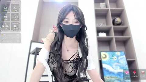 Media: Video of an East Asian woman with long black hair, wearing a black face mask, black lace top, and white off-shoulder blouse, in a modern, white room with a computer screen and shelves in the background.