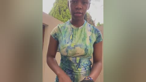 Media: Video of a young Black woman with medium-brown skin, wearing a tie-dye green and blue short-sleeved top and holding a blue bracelet. Background shows a beige wall and green foliage.