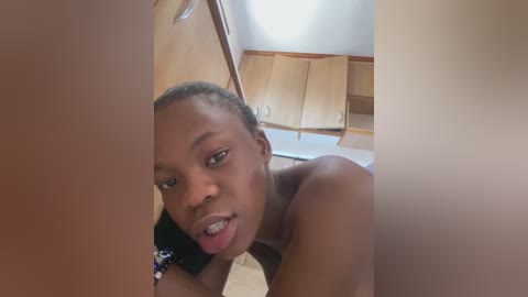 Media: Video of a young, dark-skinned African girl with braided hair, topless, licking her lips in a playful manner, set against a wooden background with cabinets.