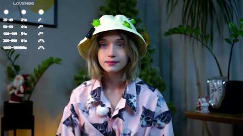 Media: Video of a young girl with shoulder-length blonde hair, wearing a white bucket hat with green leaves and a pink floral kimono, standing indoors with potted plants and a television displaying a menu screen.