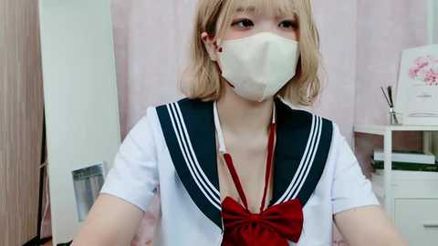 Media: Video of a young Asian woman with short blonde hair, wearing a traditional Japanese school uniform with a red bow and face mask, in a brightly lit room with light pink curtains and a white desk.