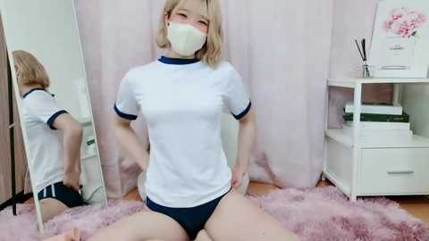 Media: Video of a petite, fair-skinned woman with blonde hair, wearing a white sports t-shirt and blue shorts, kneeling on a pink shag rug in a pink room with a mirror and white shelves.