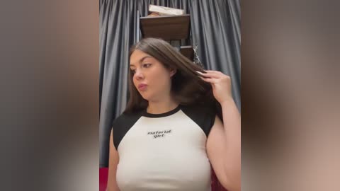 Media: Video of a young woman with long brown hair, wearing a black and white crop top, standing in front of a gray curtain backdrop. She adjusts her hair with her right hand.