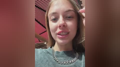 Media: Video of a young Caucasian woman with light skin, straight brown hair, and clear braces, smiling. She wears a grey sweater and a silver chain necklace. Background shows a pink room with a red curtain and wooden furniture.