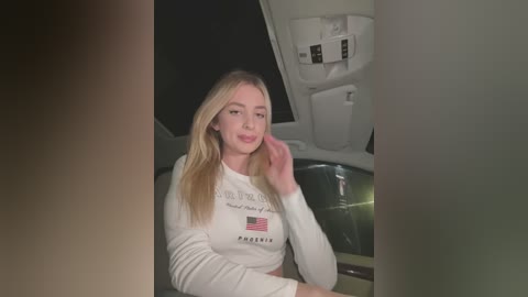 Media: A video of a blonde woman with long hair, wearing a white shirt with \"PRESIDENT\" printed on it, sitting in a car at night. The background shows a dark, shadowy interior.