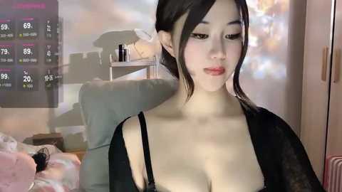 Media: Video of a young Asian woman with light skin and black hair, wearing a black bra, sitting in a dimly lit room. Background shows a TV displaying a weather forecast and a cluttered shelf.