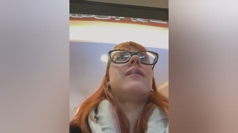 Media: Video of a young woman with red hair, wearing black-rimmed glasses and a white sweater, standing in a brightly lit room with a blurry background.