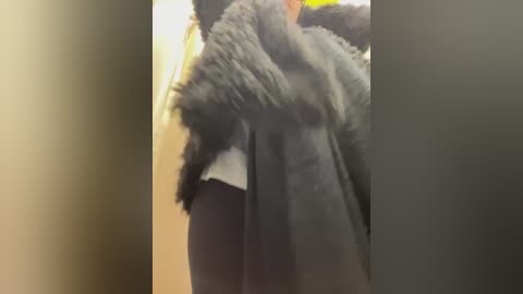 Media: A video of a person wearing a dark, fluffy fur coat, with a blurred background showing a beige wall and door frame. The coat is draped over the person\u2019s shoulders, partially covering their body.