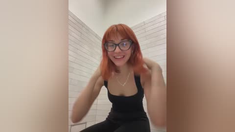 Media: Video of a young woman with shoulder-length, bright orange hair, wearing black-framed glasses and a black tank top, squatting in a small, tiled bathroom with white walls. She has a playful smile.