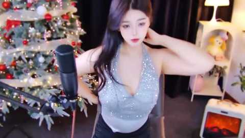 Media: Video of an East Asian woman with long black hair, wearing a glittery silver halter top and black pants, standing in a festive room with a decorated Christmas tree and fireplace.