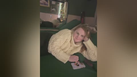 Media: A video of a young woman with fair skin, light brown hair, and freckles, wearing a cream knitted sweater, lying on a green couch in a dimly lit living room.