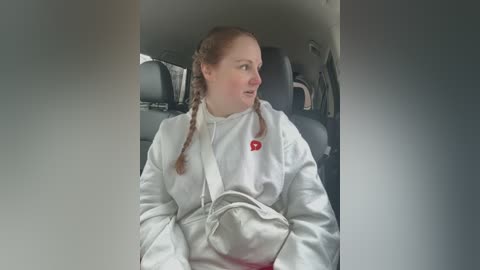 Media: Video of a white woman with light brown hair in two braids, wearing a white tracksuit, sitting in a car's backseat, looking to the side.