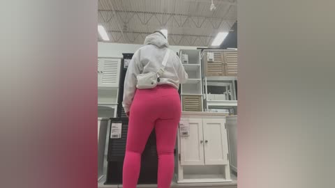 Media: Video of a person from behind in a retail store, wearing a gray hoodie and bright pink leggings, standing in front of white shelves with various boxes.