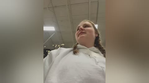 Media: Video of a young woman with auburn hair in braids, wearing a white lab coat, looking upward in a brightly lit, industrial setting with white ceilings and fluorescent lights.