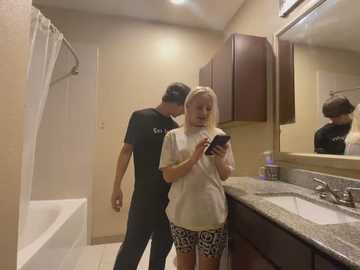 Media: Video of a blonde woman in a white shirt and patterned shorts, holding a smartphone, standing beside a man in a black t-shirt, in a beige-tiled bathroom with a granite countertop and mirror.