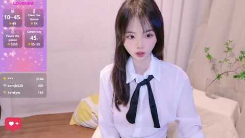 Media: Video of a young East Asian woman with long black hair, wearing a white blouse with a black bow, sitting on a bed. Background features a green plant and a digital social media interface.