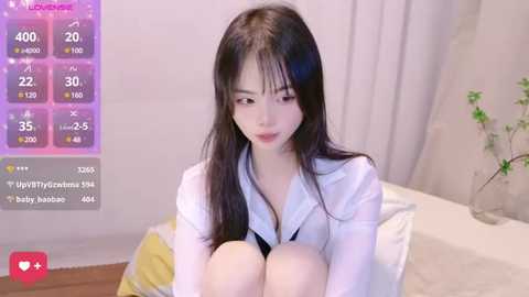 Media: A video of an East Asian woman with long black hair, fair skin, and a slim figure, wearing a white shirt, sitting on a bed. Background features a green plant, soft lighting, and a purple digital overlay with stats.