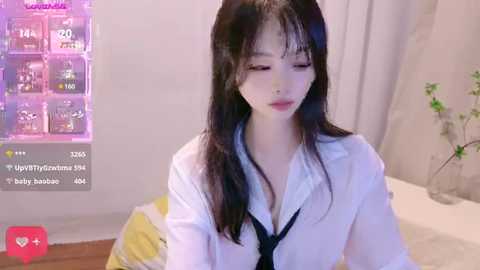 Media: Video of an Asian woman with long black hair, wearing a white shirt and black tie, standing in a bedroom with a bed, white curtains, and a poster in the background.