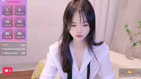 Media: A video of a young East Asian woman with long black hair and fair skin, wearing a white blouse, in a modern bedroom with a digital clock, a plant, and a bed.