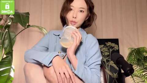 Media: Video of an Asian woman with shoulder-length brown hair, wearing a light blue shirt, sipping from a clear glass, surrounded by green plants and wooden walls.