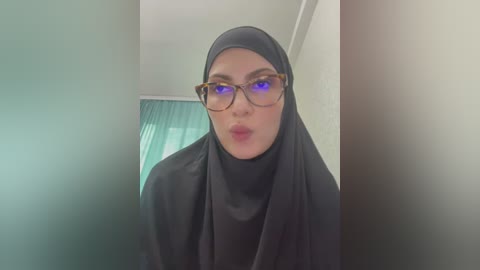 Media: A video of a light-skinned woman with large, round glasses, wearing a black hijab and a black headscarf, puckering her lips. She stands in a dimly lit room with a teal curtain in the background.