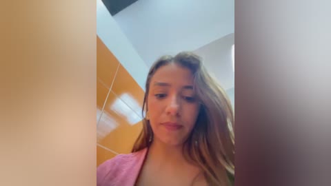 Media: A video of a young Caucasian woman with light skin and long brown hair, wearing a pink towel, standing in a tiled bathroom with orange walls and white ceiling.