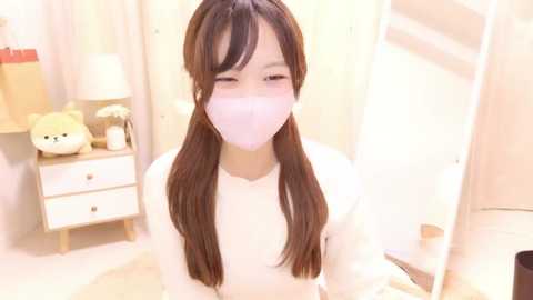 Media: Video of a young Asian woman with long brown hair, wearing a white mask, sitting in a brightly lit, minimalist bedroom with beige walls, a wooden dresser, and a stuffed animal.