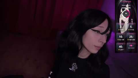 Video of a young woman with long, wavy black hair and pale skin, wearing a black outfit, standing in a dimly lit room with purple lighting. She appears to be in a gaming context, with a digital overlay showing stats.