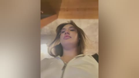 Media: A video of a young woman with light skin and straight, shoulder-length brown hair, wearing a white zip-up hoodie, standing in a dimly lit room with wooden floors and a white rug.