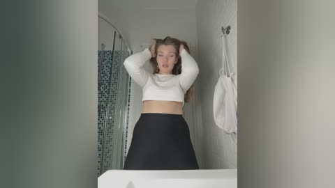 Media: Video of a young woman with fair skin, long brown hair, wearing a white crop top and black high-waisted pants, posing in a modern bathroom with a white towel and mosaic-tiled shower.