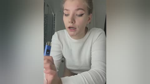 Media: Video of a young Caucasian woman with light skin, blonde hair tied back, wearing a white long-sleeve shirt, applying mascara with a blue wand, in a bathroom with a light-colored wall and showerhead.