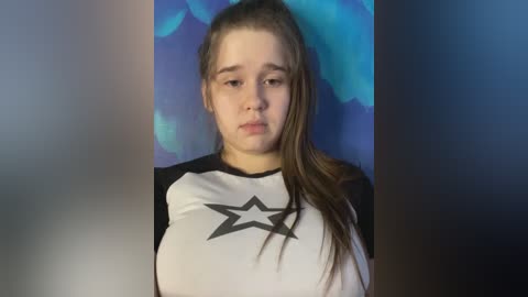 Media: A video of a young Caucasian girl with fair skin and long brown hair, wearing a black-and-white star-printed shirt, against a blue abstract background. She has a neutral expression.