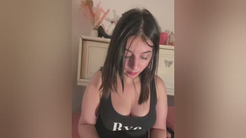 Media: A video of a young woman with long, dark hair wearing a low-cut black tank top, sitting in a cozy, dimly lit room with beige walls and a floral arrangement.