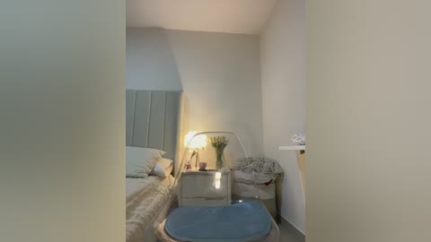 Media: A video of a small, minimalist bedroom with a beige wall, a bed with a light blue headboard, a nightstand with a lamp, and a blue cushioned stool.