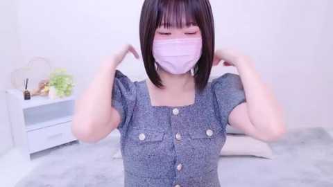 Media: A video of an East Asian woman with straight black hair and bangs, wearing a light purple mask and a blue button-up shirt. She's adjusting her mask in a minimalist white room with a white shelf and plants.