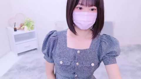 Media: Video of an Asian woman with straight, shoulder-length black hair and light skin, wearing a blue short-sleeve dress with puffed shoulders, a white face mask, and a white headband. She stands in a minimalist, white room with a white nightstand and green plants in the background.