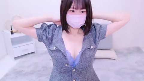 Media: A video of an Asian woman with short dark hair, wearing a gray button-up dress with a lace bra, a face mask, and a bracelet, standing in a minimalist white room with a bed and a nightstand.