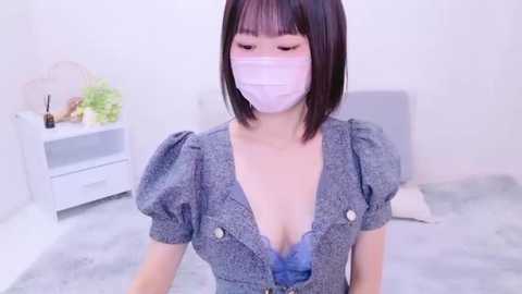 Media: Video of an East Asian woman with straight, shoulder-length black hair, wearing a blue plaid dress with a deep V-neckline, revealing cleavage, and a white face mask. Background includes a white bedside table with a green plant and a white cushion.