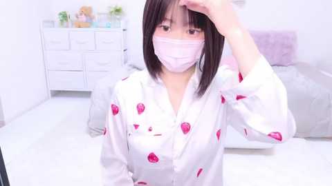 Media: Video of an Asian woman with straight black hair, wearing a white robe with pink strawberry prints, a face mask, and a white hat, standing in a white bedroom with a bed, dresser, and potted plants.