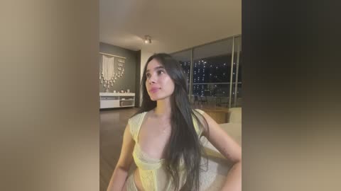 Media: Video of a young, slender woman with long, straight black hair, wearing a sheer yellow dress, standing indoors, gazing at a cityscape through large windows.