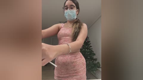 Media: A video shows a young woman with light brown skin, long brown hair, and glasses, wearing a pink floral dress and a face mask, cleaning a window. A Christmas tree is visible in the background.