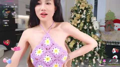 Media: Video of a slim, Asian woman with long black hair, wearing a pink crochet halter top with white daisy print, posing indoors near a decorated Christmas tree.