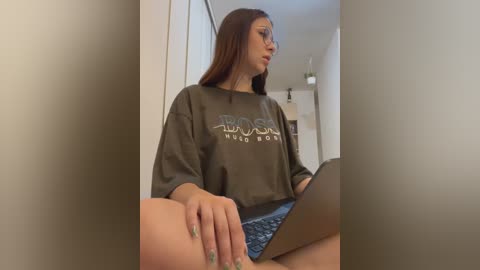 Media: A video of a young woman with long brown hair, wearing a loose, dark green sweatshirt and denim shorts, sitting on a bed with a laptop in front of her.