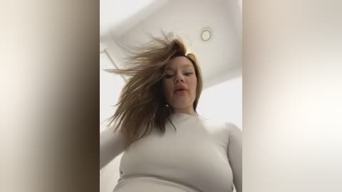 Media: Video of a woman with light skin and long, messy brown hair, wearing a white long-sleeve top, captured from a low angle, emphasizing her facial features and hair movement.