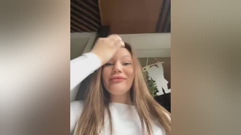 Media: Video of a young woman with long, straight, light brown hair, wearing a white long-sleeve shirt. She's smiling, her right hand touching her head, in a cozy, well-lit room with wooden beams and a potted plant.