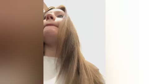 Media: A video showing a close-up of a young woman with long, straight brown hair, fair skin, and a light complexion. The background is a plain, light-colored wall. The image captures her slightly tilted head and a portion of her face, emphasizing a serene expression.