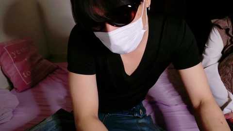 Media: Video of a person with long black hair, wearing a black shirt, white mask, and blue jeans, seated on a pink bed, reading a book.