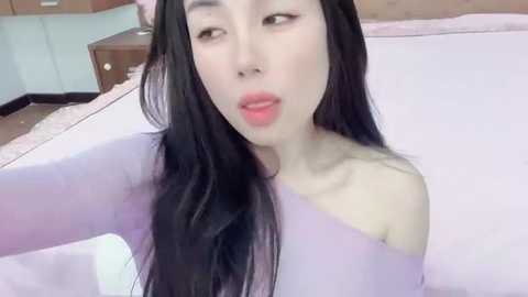 Media: A video of a young Asian woman with long black hair, wearing a light purple off-shoulder top, in a bedroom with white bed linens and wooden furniture.